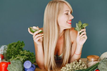 A smiling woman holding avocado and basil, surrounded by fresh vegetables and fruits for healthy eating.